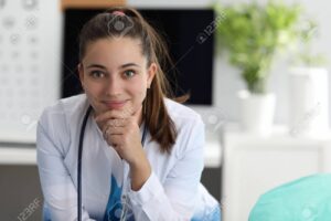 Portrait of beautiful young female doctor sitting in cabinet in hospital. Woman wearing medical gown and professional equipment stethoscope on neck. Medicine concept